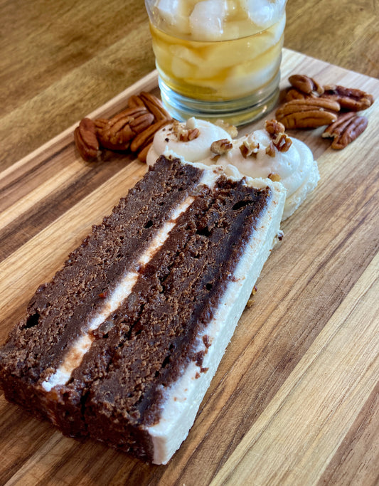 Slice of chocolate cake with white frosting on a wooden cutting board with pecans and a glass of iced tea.